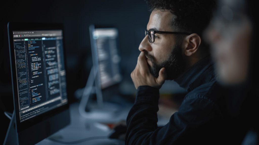 man with glasses and a beard looking thoughtfully at a computer screen with code