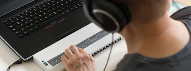 A person with vision loss, wearing headphones, who is using a computer which has a braille output pad